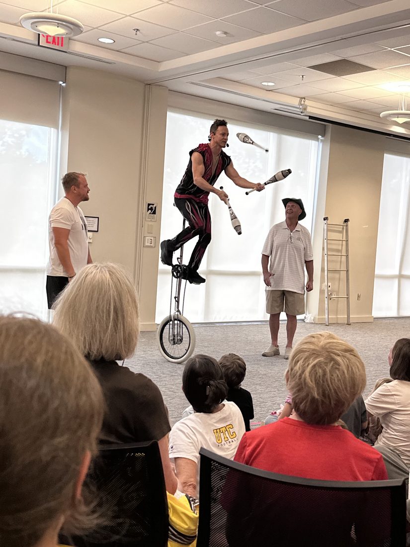 Rocco juggling on a tall unicycle while a volunteer helps in a library program room